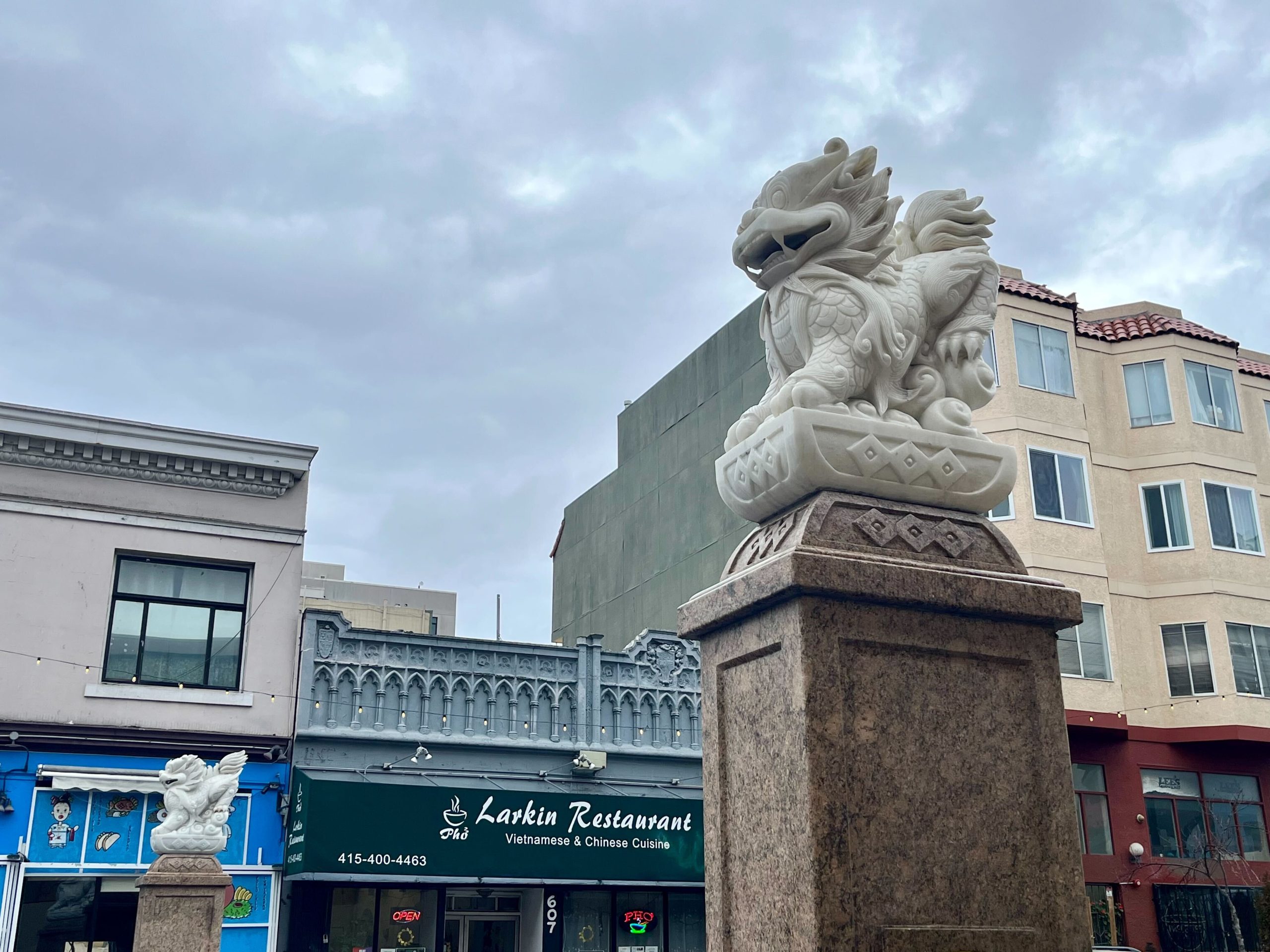 Marble dragon statues atop stone pedestals stand in front of storefronts, including Larkin Restaurant, under a cloudy sky.