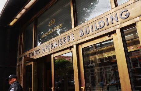 Exterior entrance of the United States Appraisers Building at 630 Sansome Street, featuring brass signage and glass doors, with an ICE sign visible as a person walks past on the left.
