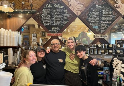 Five people stand smiling behind a coffee shop counter, with menu boards, cups, and coffee products visible in the background.