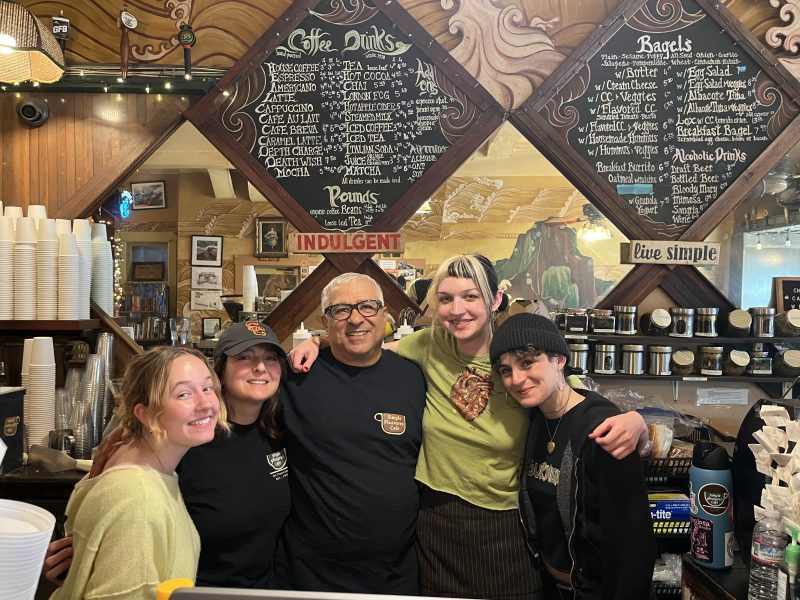 Five people stand smiling behind a coffee shop counter, with menu boards, cups, and coffee products visible in the background.