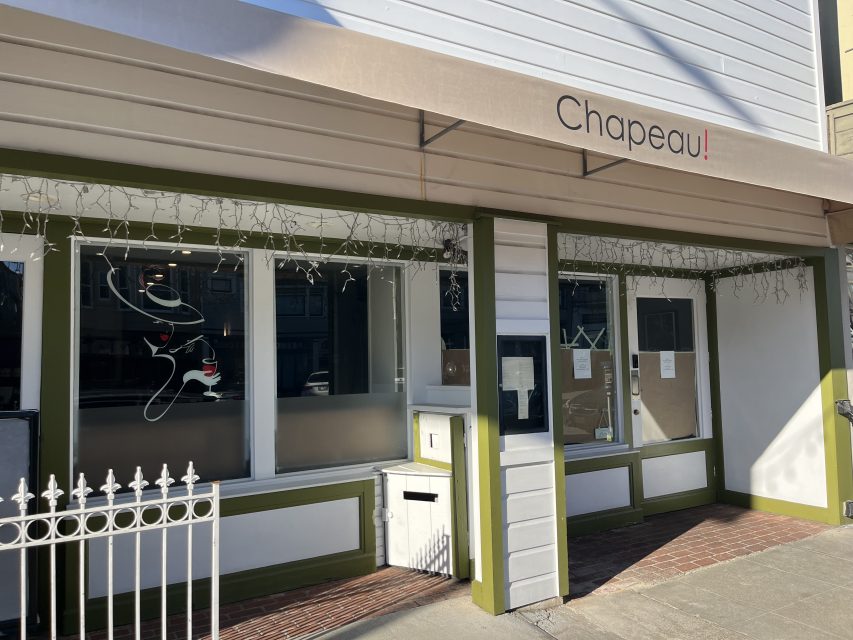 Storefront of Chapeau! restaurant with a beige awning, green trim, window art of a face with a hat, and papers posted on the door.