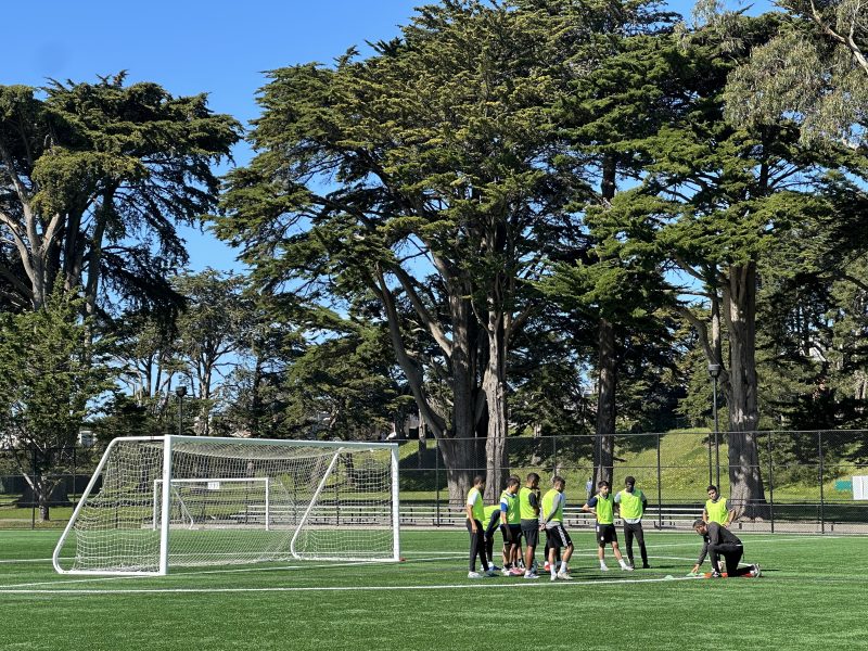 A group of people in green vests gathers near a soccer goal on an outdoor field surrounded by trees under a clear blue sky.