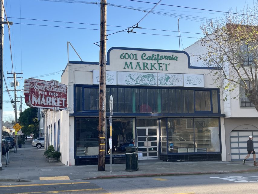 Street view of a corner market with “6001 California Market” and “Apple Delecta Fine Food Market” signs, large front windows, and a tree to the right of the building.