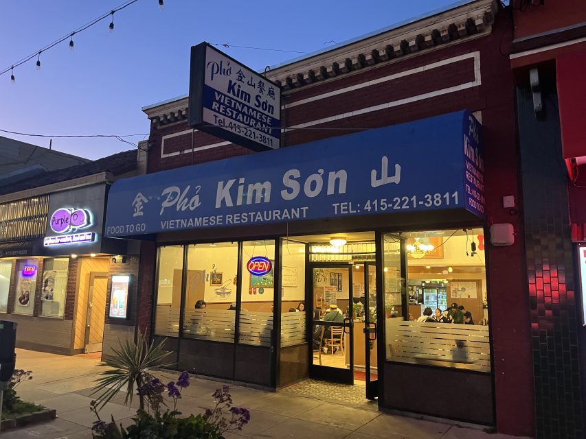 Storefront of Pho Kim Son Vietnamese Restaurant at dusk, with people dining inside and blue signage above the entrance displaying the restaurant's name and phone number.