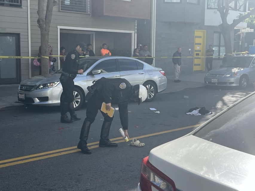 Police officers examine items on a city street behind yellow tape, with bystanders watching and cars parked nearby in daylight.