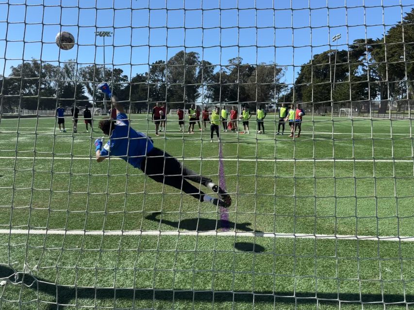 A soccer goalkeeper in blue dives to block a ball during a penalty kick, with players watching from the field behind the net.