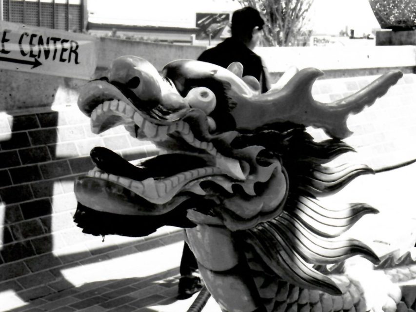 A close-up of a dragon-shaped decorative statue outdoors, with a person walking in the background and a partial sign visible on the left.