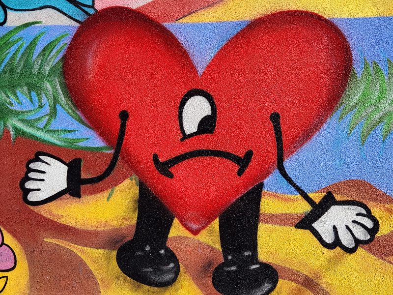 A mural of a large red heart with arms, legs, gloved hands, and a sad face stands against a colorful outdoor background.
