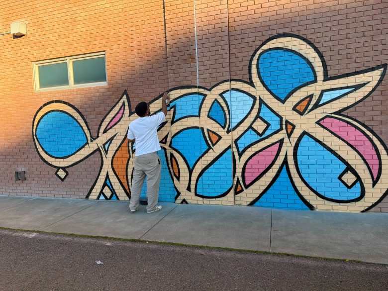A person in a white shirt and gray pants is spray painting colorful abstract graffiti on a brick wall.