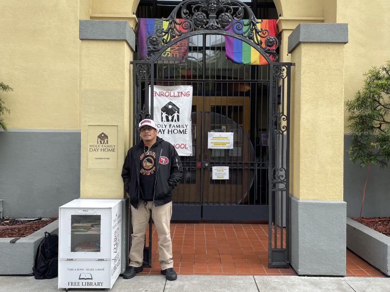 A man stands in front of a gated entrance to a building with banners, rainbow flags, and a free library box nearby.