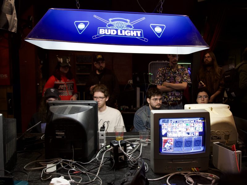 People play video games on old CRT monitors at a crowded event, with a Bud Light sign hanging overhead and game consoles and wires visible on the table.