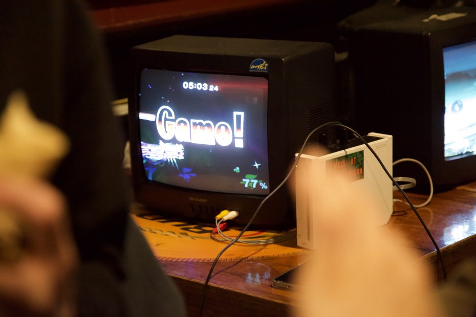 A CRT monitor on a wooden table displays a "Game!" screen from a video game, connected to a white console with cables.