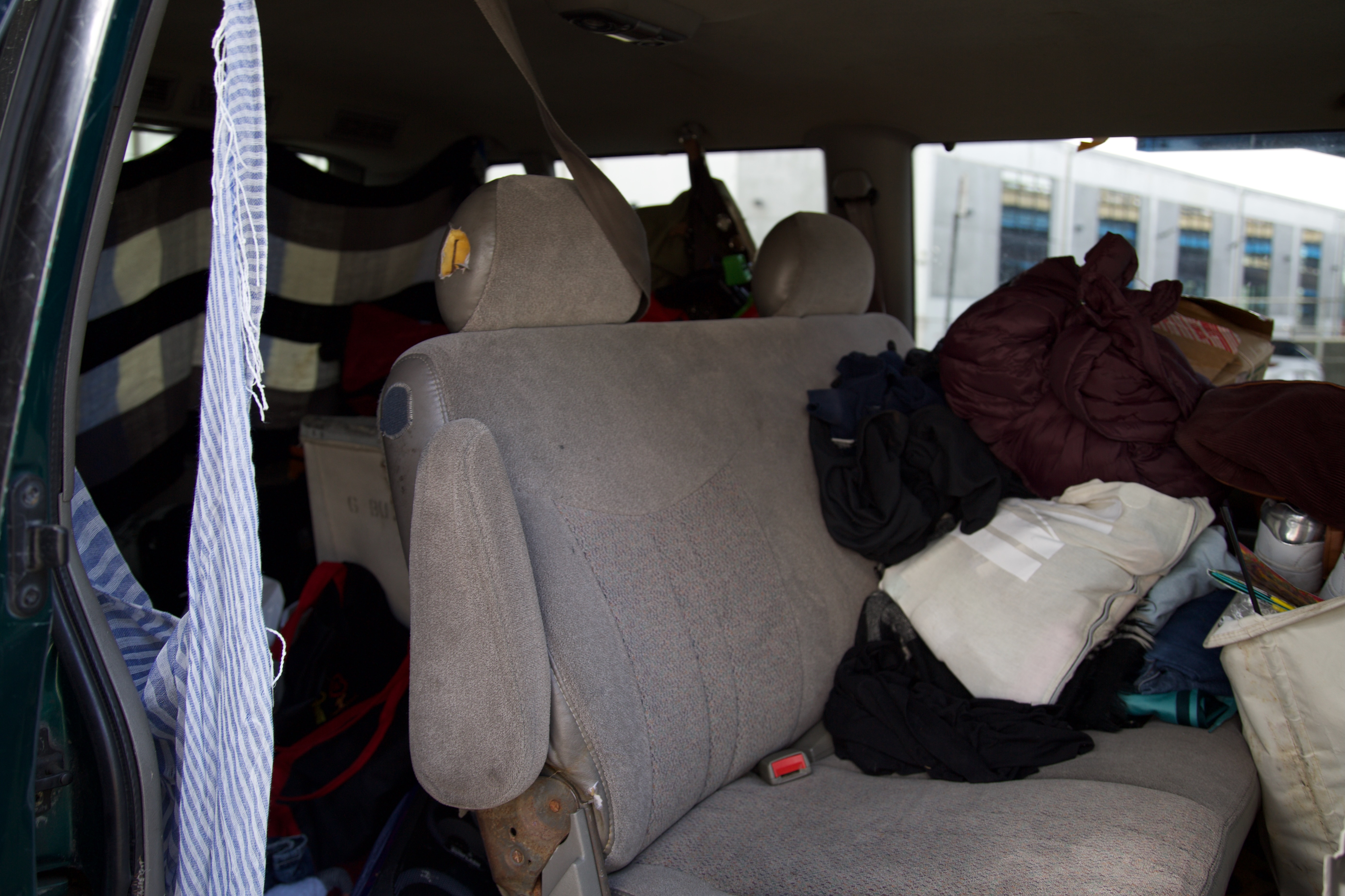 The interior of a car with the back door open, showing a cluttered backseat filled with clothes, bags, and other personal items.