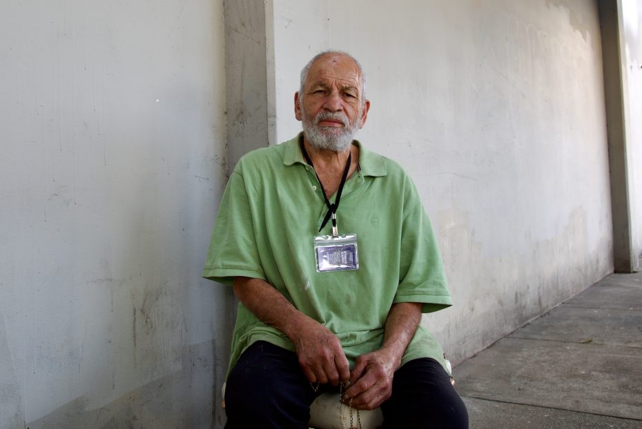 An older man with a beard sits on a stool outside against a blank wall, wearing a green polo shirt and a visible lanyard with an ID badge.