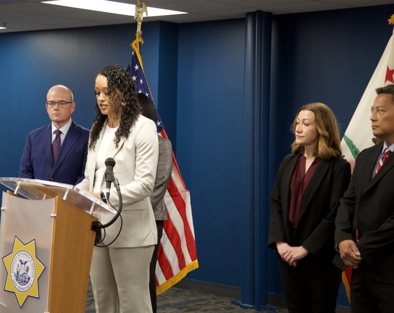 A woman speaks at a podium with four people beside her in a Davis conference room, framed by American and California flags.