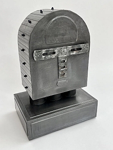 A metallic, abstract sculpture resembling a simplified human face with rectangular features, bolts on the sides, and mounted on a rectangular base.