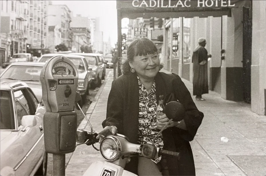 A woman sits on a scooter parked by a meter on a city street near the Cadillac Hotel, smiling and holding a microphone. Cars line the street in the background.