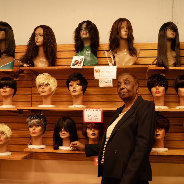 An older woman stands in front of a display of wigs on mannequins in a store, with sale signs and a no returns notice visible.