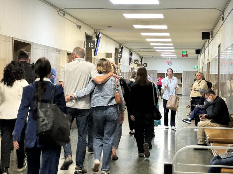 People walk down a hallway with numbered signs overhead, some sitting on benches along the side. A couple walks arm-in-arm among others moving in both directions.