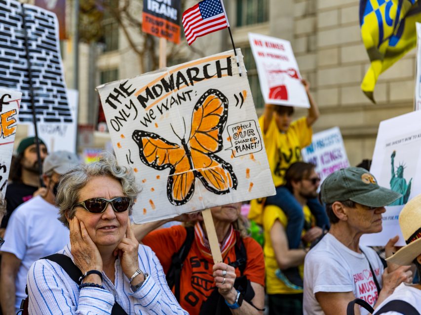 A group of protesters at a rally, one holding a sign with an orange monarch butterfly and the text "The Only Monarch We Want: Protect Our Planet.