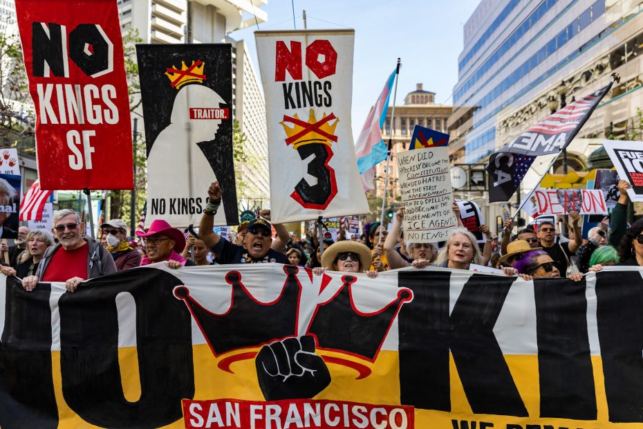 A group of protesters march in San Francisco holding banners and signs reading “NO KINGS SF” and “NO KINGS,” with crown graphics and a large protest banner in the foreground.