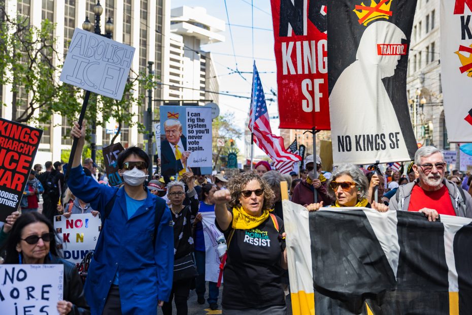 Protesters march in a city holding signs and banners, including messages like "Abolish ICE," “No Kings SF,” and “Resist,” with American flags in the background.