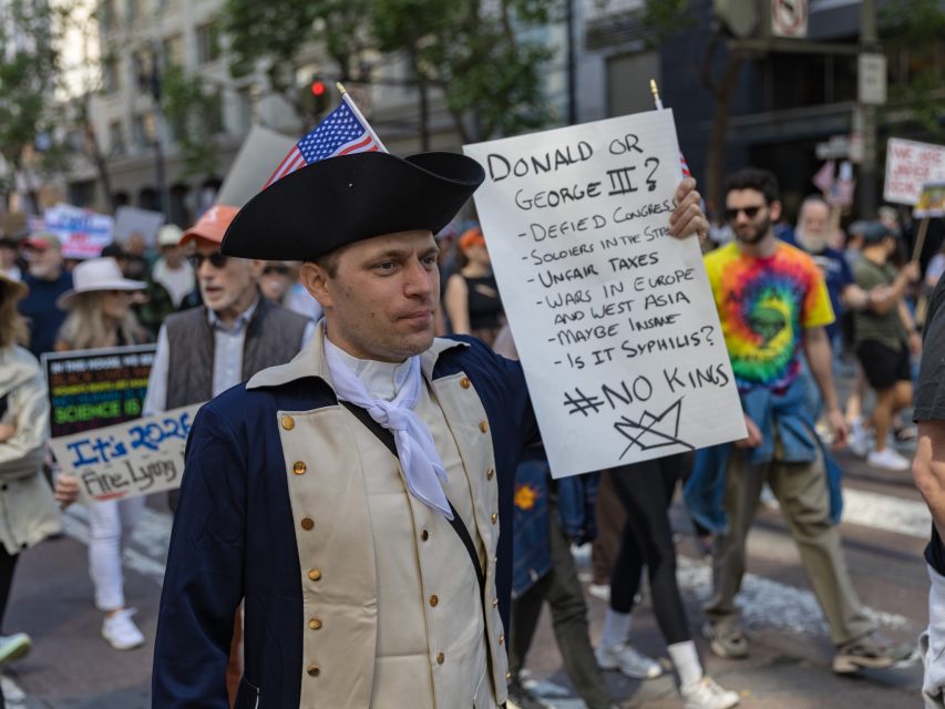 A person in colonial attire holds a sign referencing Donald Trump and George III at a protest; other people with signs walk in the background on a city street.