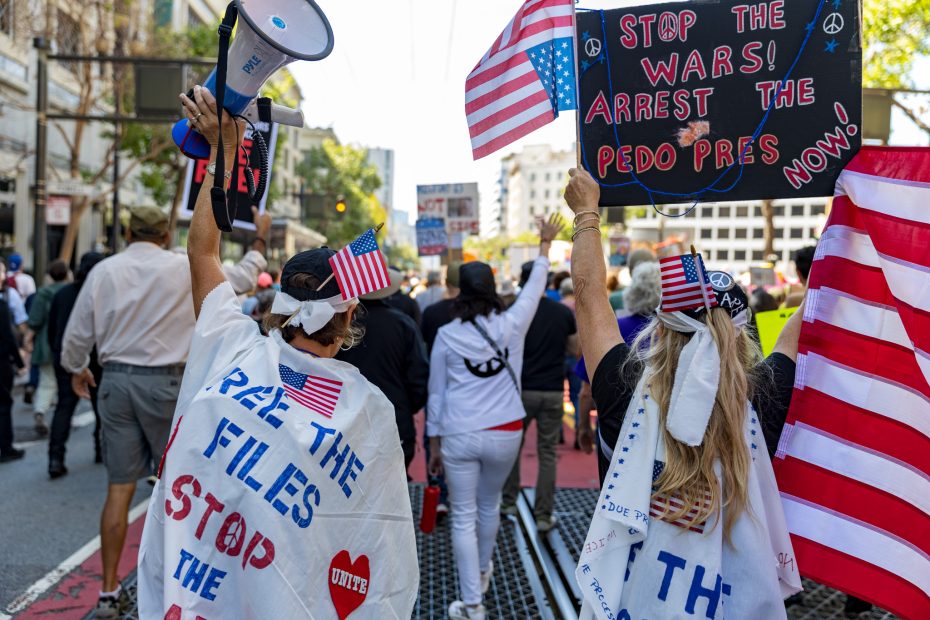 Two people draped in flags and signs protest on a city street among a crowd, holding up posters with anti-government and anti-war messages.