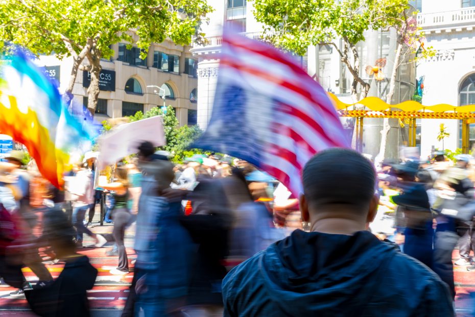 A crowd of people move along a city street during a daytime protest, with an American flag and a rainbow flag visible in motion blur.