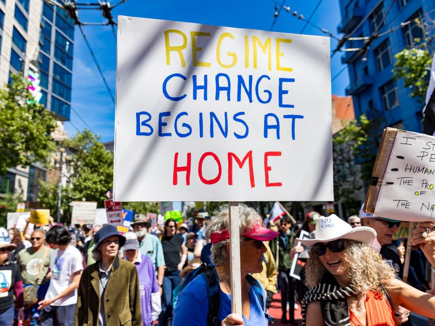 A group of people march outdoors holding signs, including one large sign that reads, "Regime change begins at home.