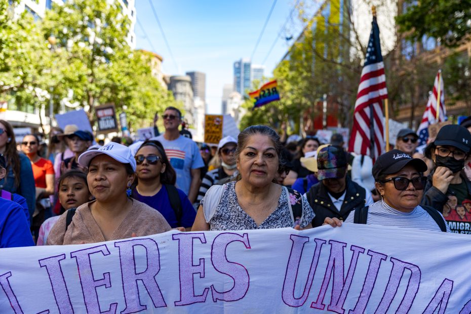 A diverse group of people march together in a city street holding a banner that reads "MUJERES UNIDAS." American and rainbow flags are visible in the background.