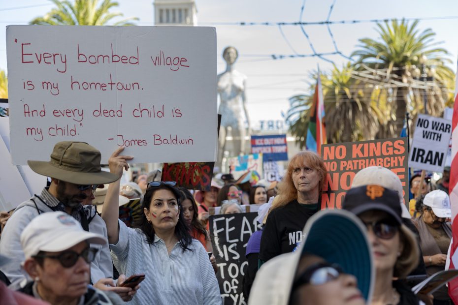 A crowd at a protest holds signs, including one quoting James Baldwin and others with messages against war and patriarchy; palm trees and buildings are visible in the background.
