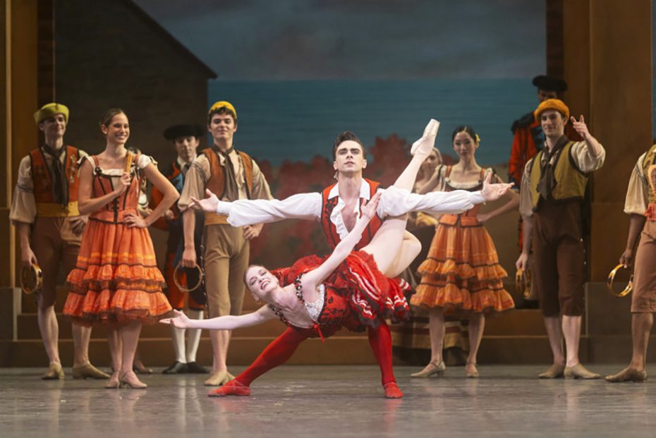 A male and female ballet dancer perform a dramatic pose center stage, surrounded by other dancers in period costumes during a ballet performance.