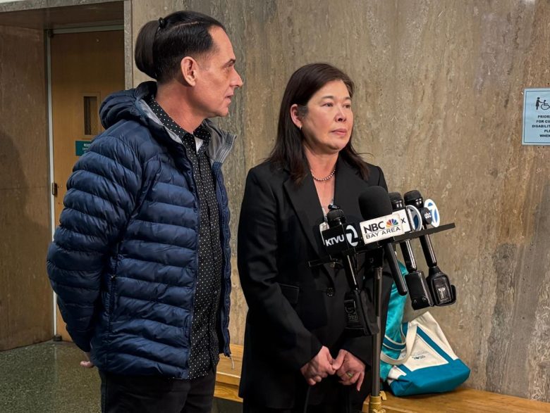 Two people stand in front of microphones during a press conference in a hallway, with a wooden bench and signs visible in the background.