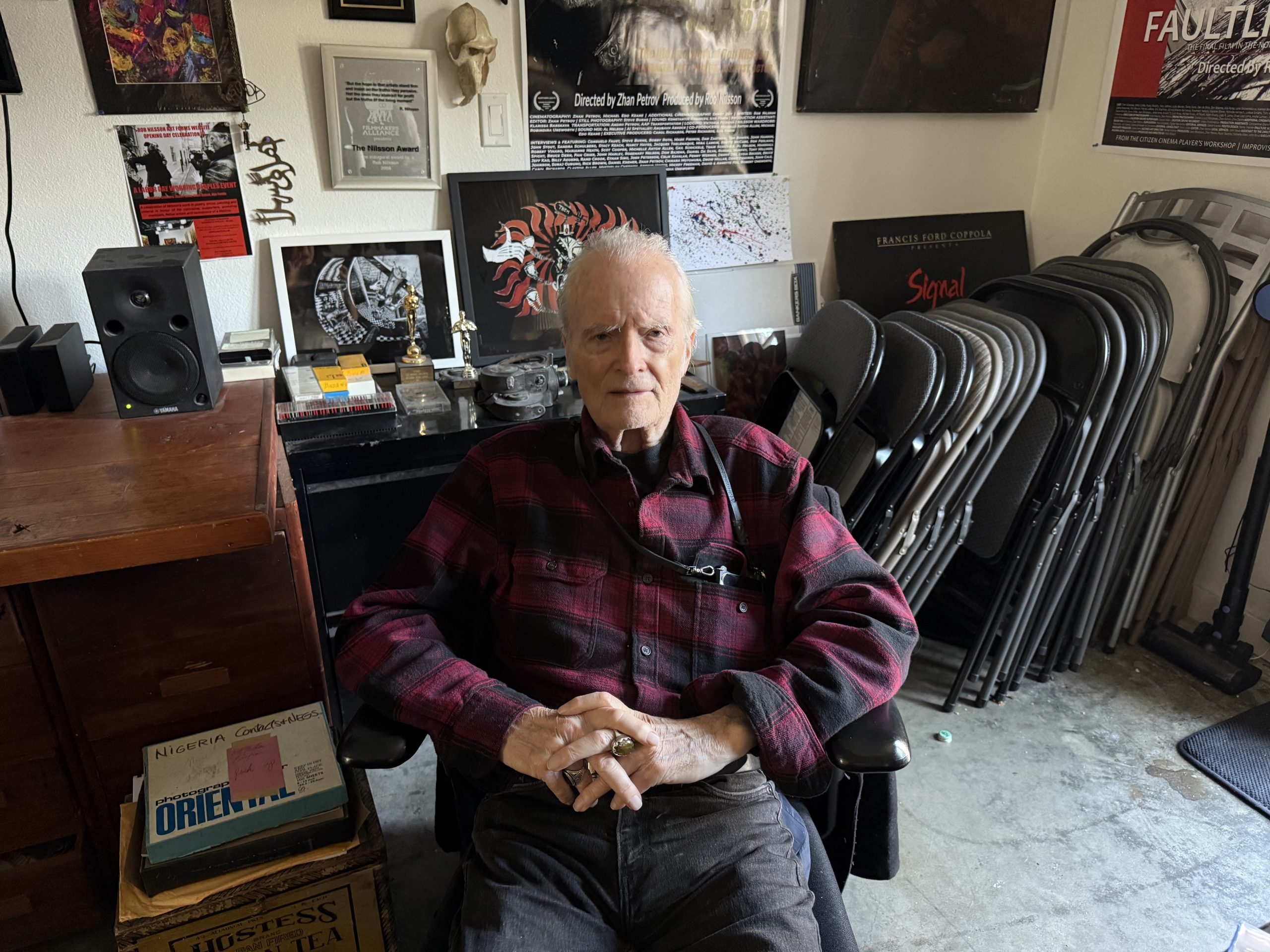 An elderly man in a red plaid shirt sits in a chair in a cluttered room, surrounded by folding chairs, art, audio equipment, and memorabilia from Nilsson in the background.