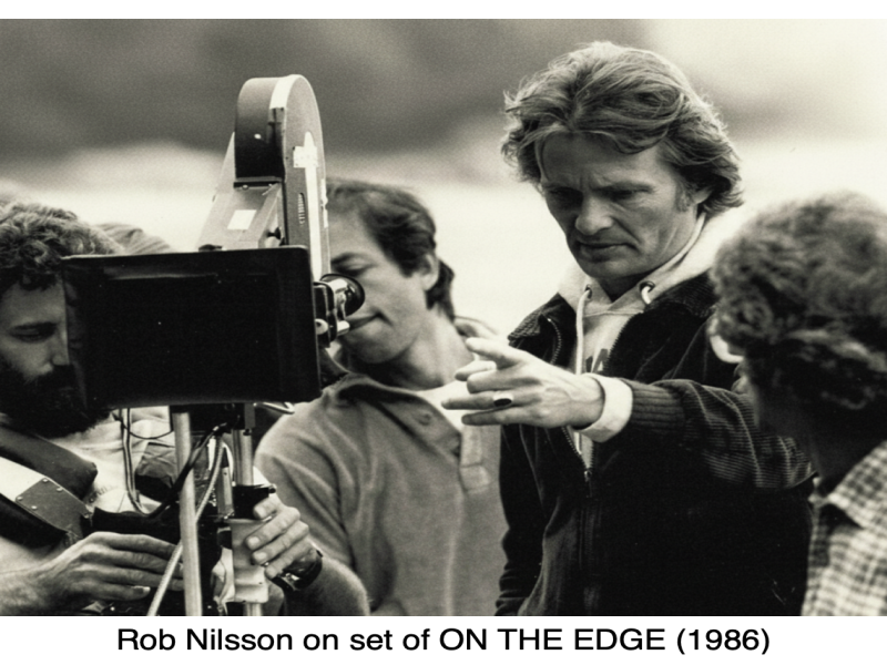 Rob Nilsson, surrounded by his crew, directs a scene with a film camera on the set of "On the Edge" (1986), showcasing Nilsson’s hands-on approach to filmmaking.