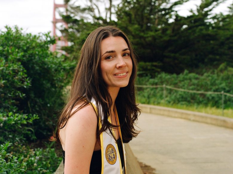 A young woman with long brown hair, wearing a graduation stole, smiles while standing on a path with greenery and a bridge structure in the background.
