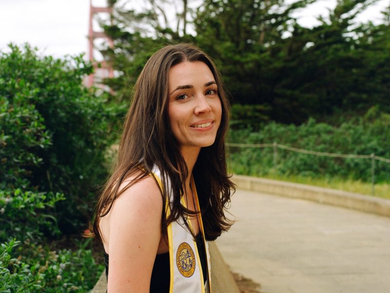 A young woman with long brown hair, wearing a graduation stole, smiles while standing on a path with greenery and a bridge structure in the background.