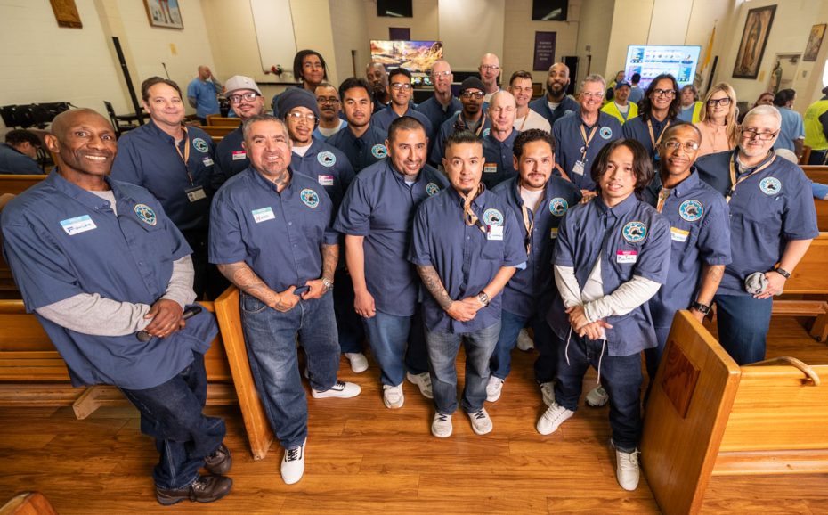 A group of people in matching blue shirts posing together inside a room with wooden benches, likely in a community or church setting.