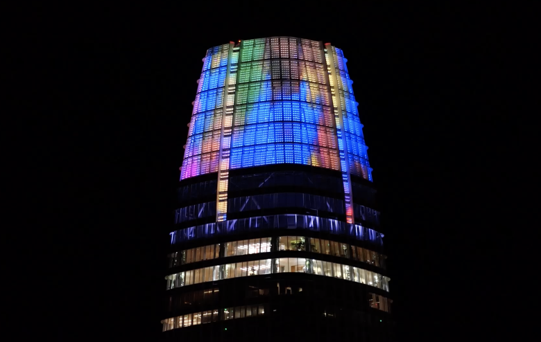 A high-rise building at night with its upper section illuminated in multicolored lights, including blue, green, pink, and yellow, against a dark sky.