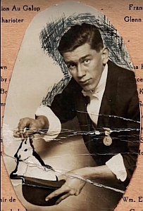 A man in formal attire holds a shoe with a small object inside, looking at the camera. The photo is old and has visible creases and damage.