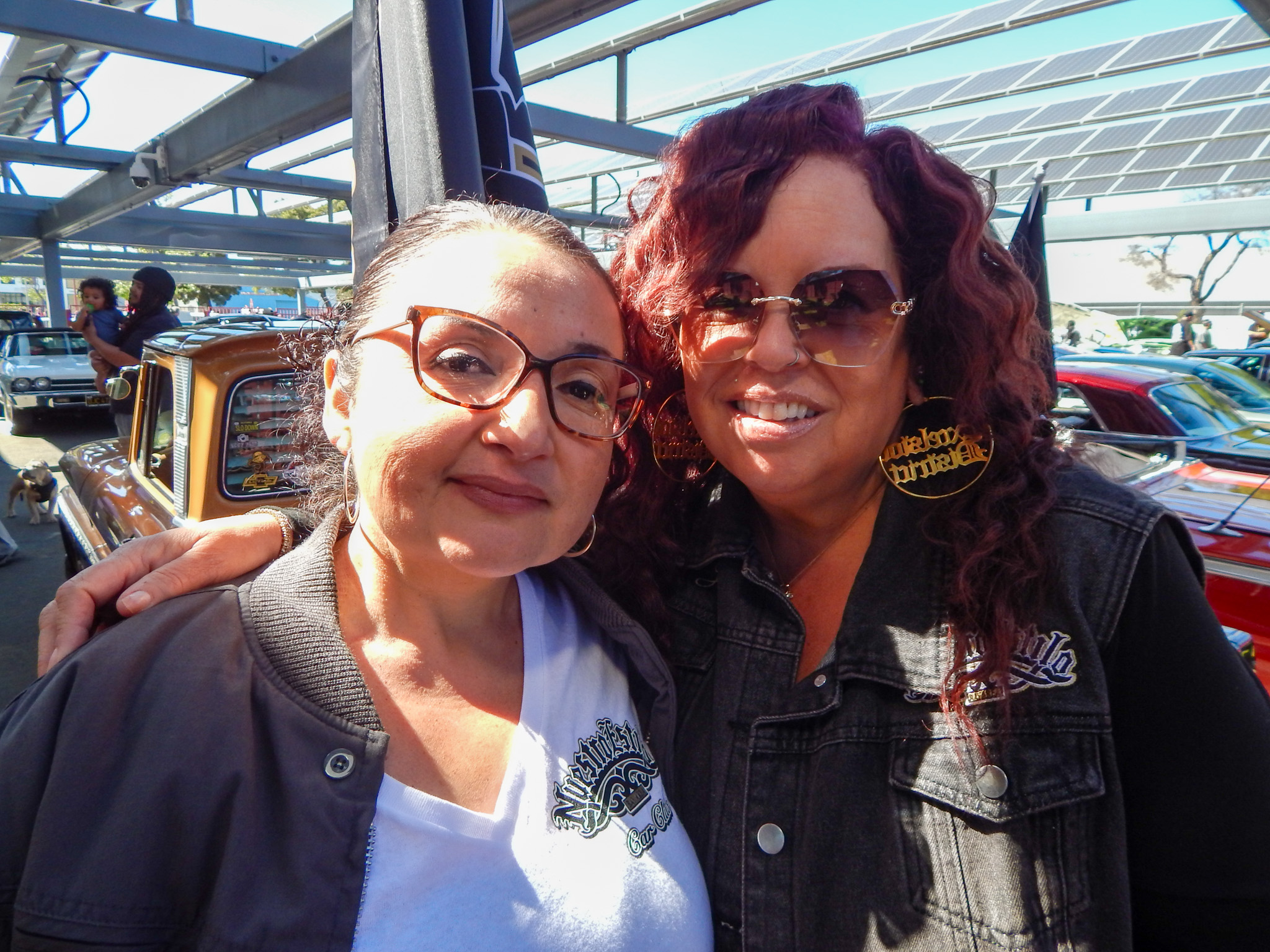 Two women wearing glasses and jackets stand close together and smile at an outdoor car show with classic cars in the background.