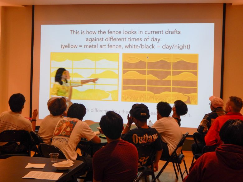 A presenter points to a projected slide showing draft designs of a metal art fence, with attendees seated and viewing the presentation.