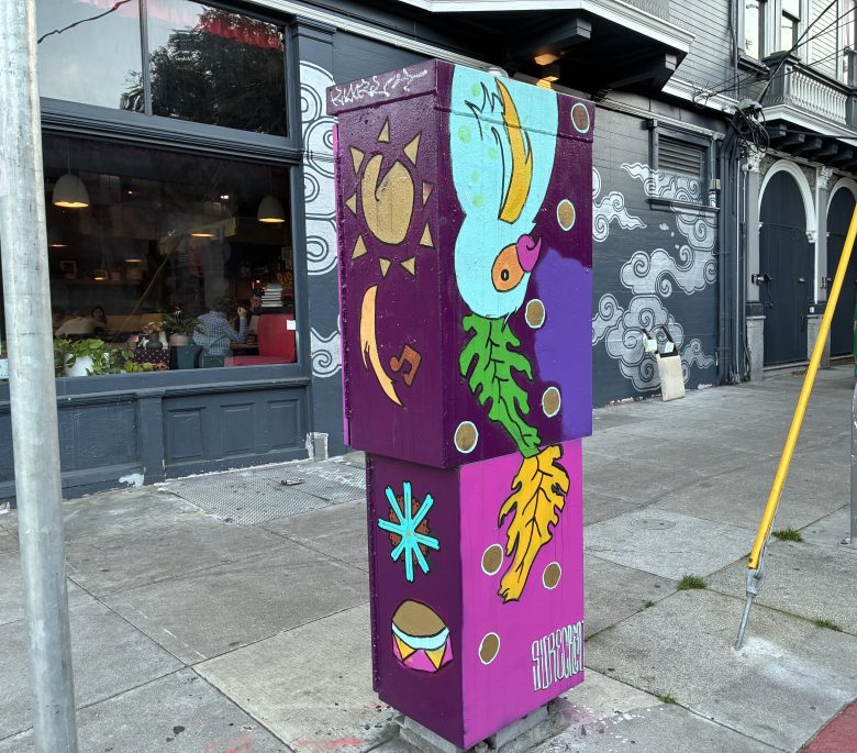 A utility box on a city sidewalk is painted with colorful abstract designs, including a leaf, sun, star, and drum, in front of a cafe and mural-covered wall.