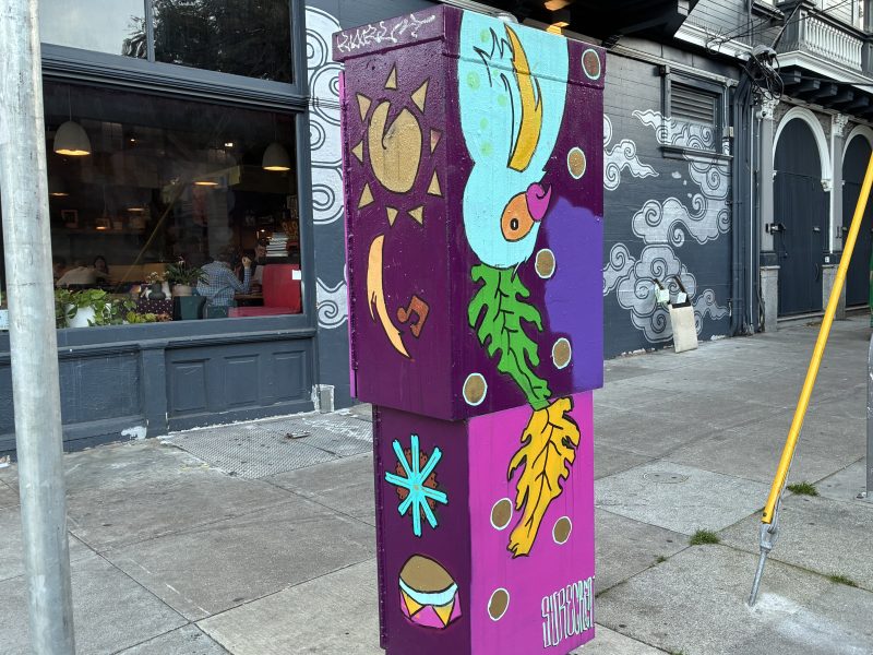 A utility box on a city sidewalk is painted with colorful abstract designs, including a leaf, sun, star, and drum, in front of a cafe and mural-covered wall.