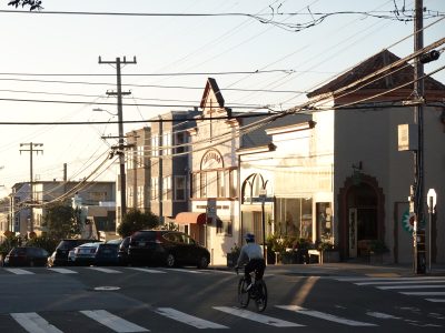 A person on a bicycle rides through a sunny intersection lined with parked cars and storefronts in a neighborhood with overhead utility wires.
