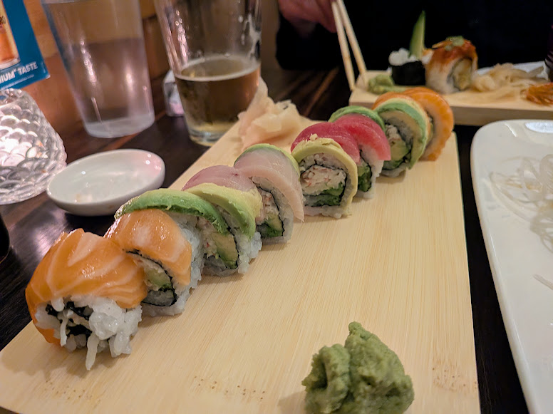 A wooden platter with assorted sushi rolls topped with avocado and various fish, accompanied by wasabi. A glass of beer and more sushi are visible in the background.