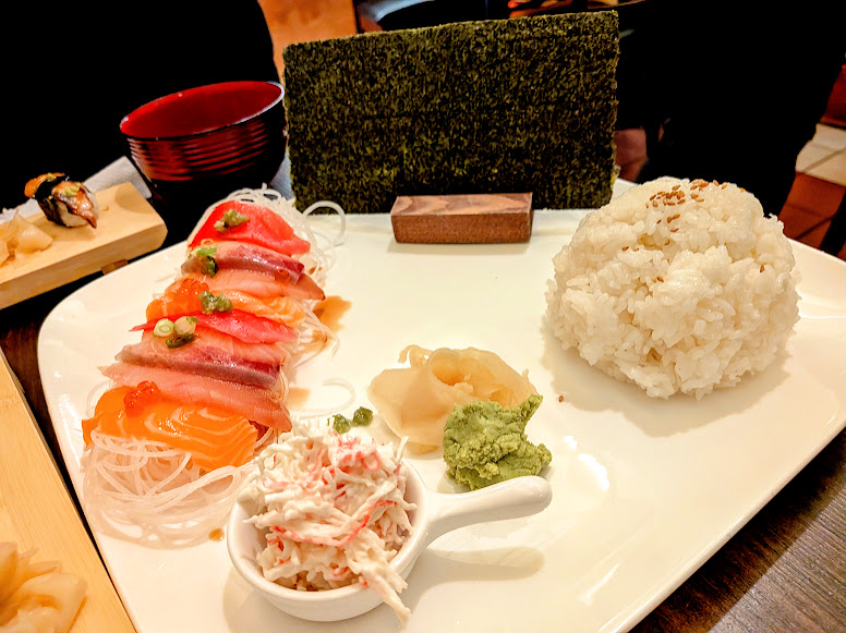 A plate with sashimi slices, shredded crab salad, pickled ginger, wasabi, a mound of white rice, seaweed sheets, and a bowl of miso soup in the background.