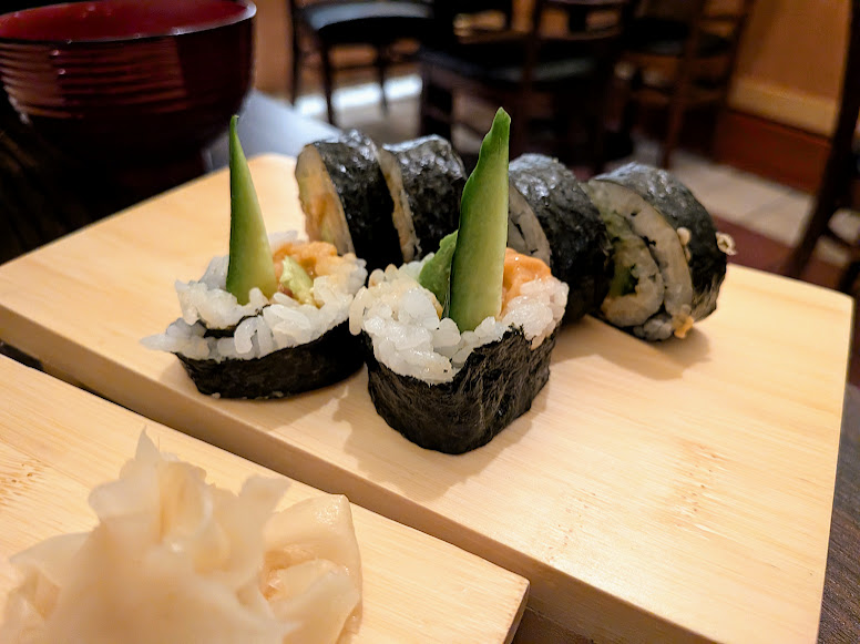 Sushi rolls filled with rice and vegetables, topped with upright asparagus, served on a wooden board with a side of pickled ginger.