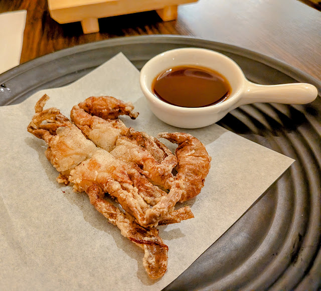 A piece of fried soft-shell crab is served on parchment paper next to a small dish of dipping sauce on a dark plate.